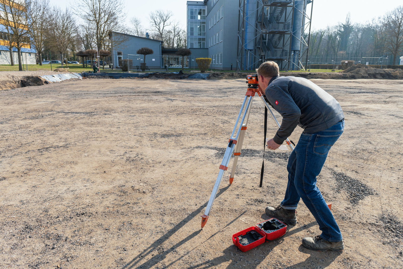 Ein Bauarbeiter nutzt ein Nivelliergerät auf einer Baustelle, um eine präzise Ausrichtung auf einer ebenen Fläche zu überprüfen. Im Vordergrund liegt eine Wasserwaage.