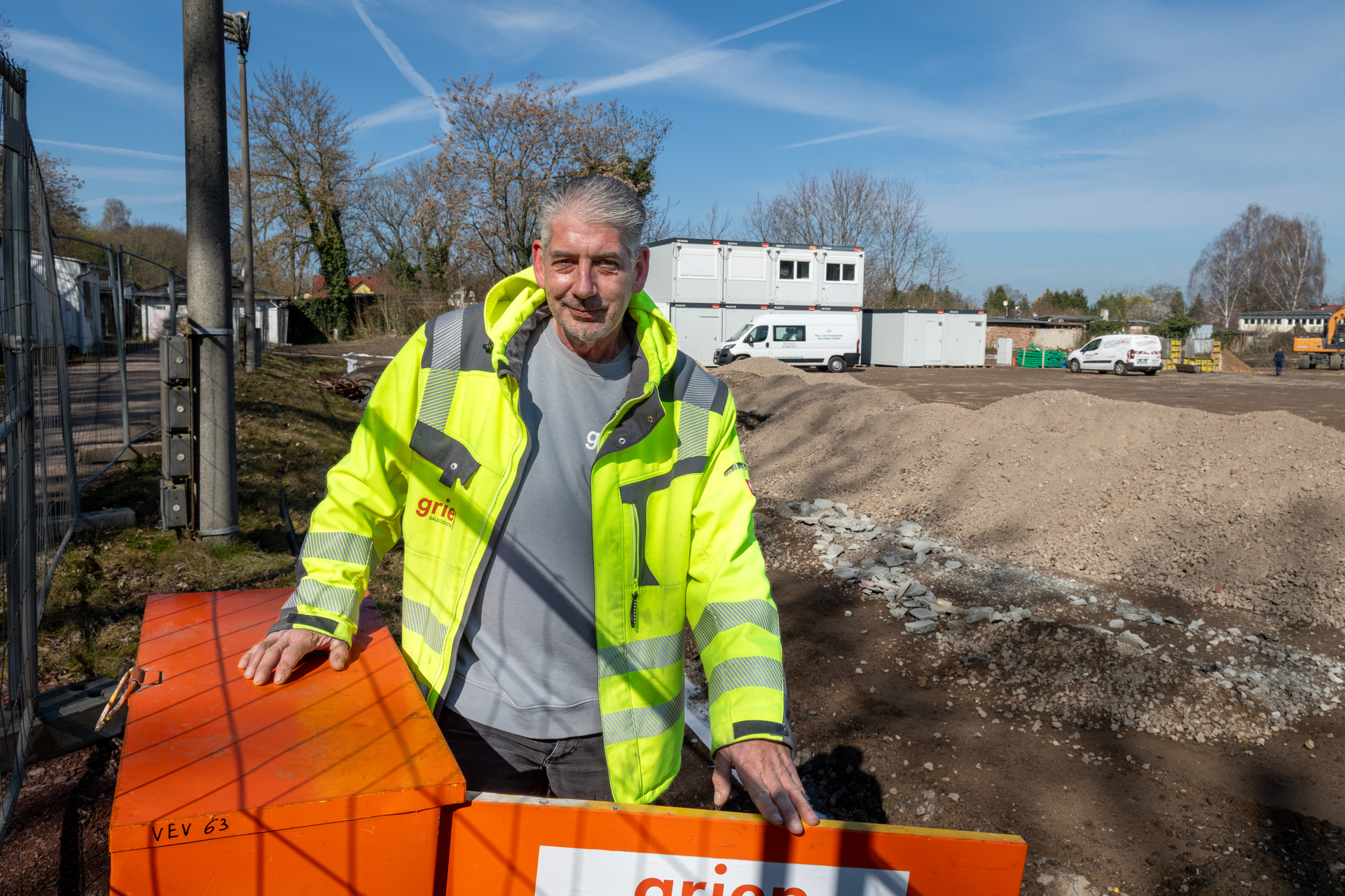 Ein Mann in gelber Warnweste mit Firmenlogo „grie“ steht lächelnd auf einer orangefarbenen Baustellenabsperrung, umgeben von einer unfertigen Baustelle mit Fahrzeugen und Gerüsten im Hintergrund.