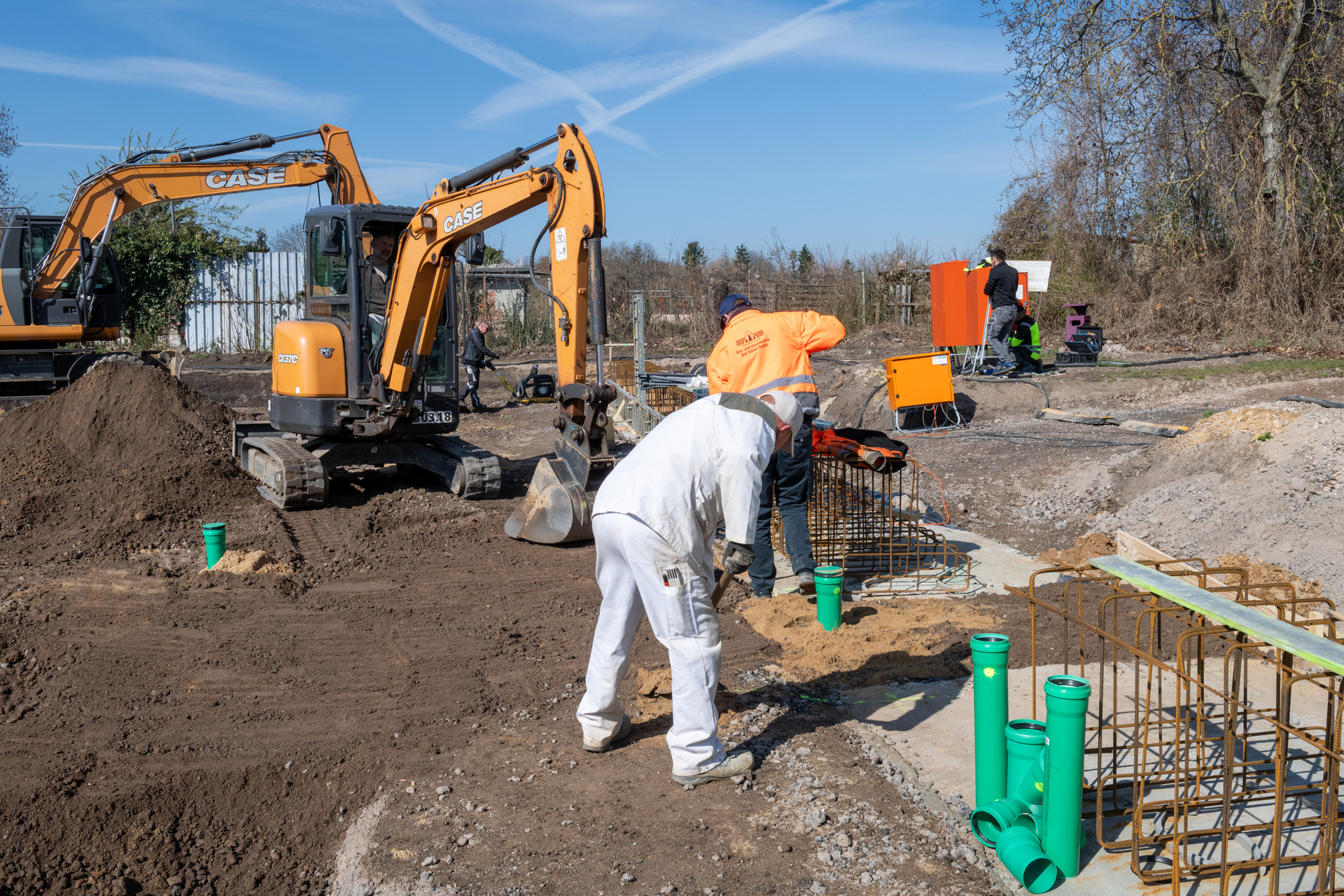 Auf einer Baustelle arbeiten zwei Personen: Eine in weißer Schutzbekleidung und Helm, die Betonrohre in ein vorbereitetes Fundament setzt, während im Hintergrund ein gelber Bagger der Marke CASE aktiv ist.