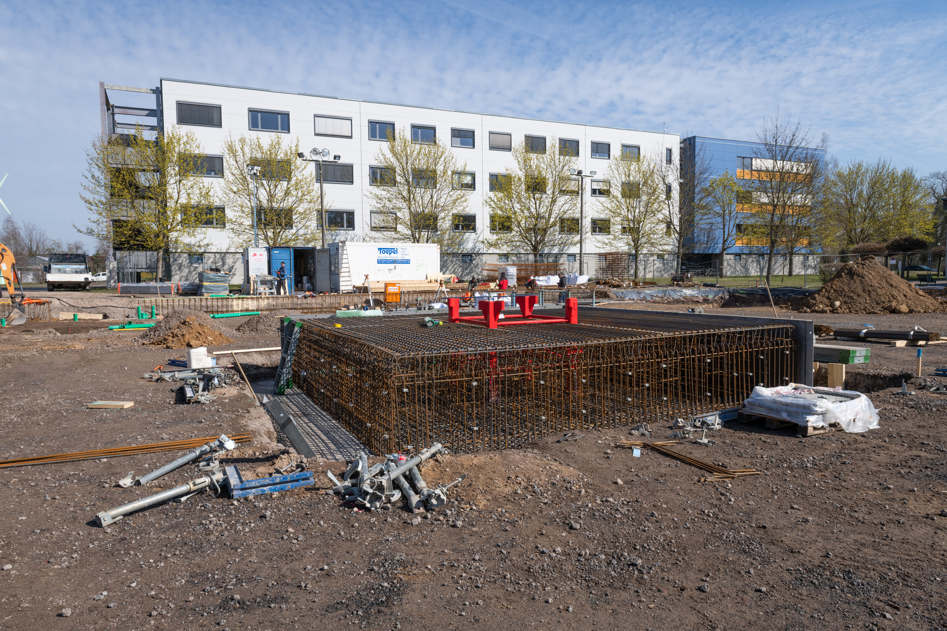 Baustelle mit vorbereitetem Stahlbeton-Grundgerüst für ein Fundament, umgeben von Baustoffen wie Stahlstangen, Schutt und Werkzeugen. Im Hintergrund ein mehrstöckiges Gebäude in Bauphase mit offenen Fenster- und Türöffnungen.