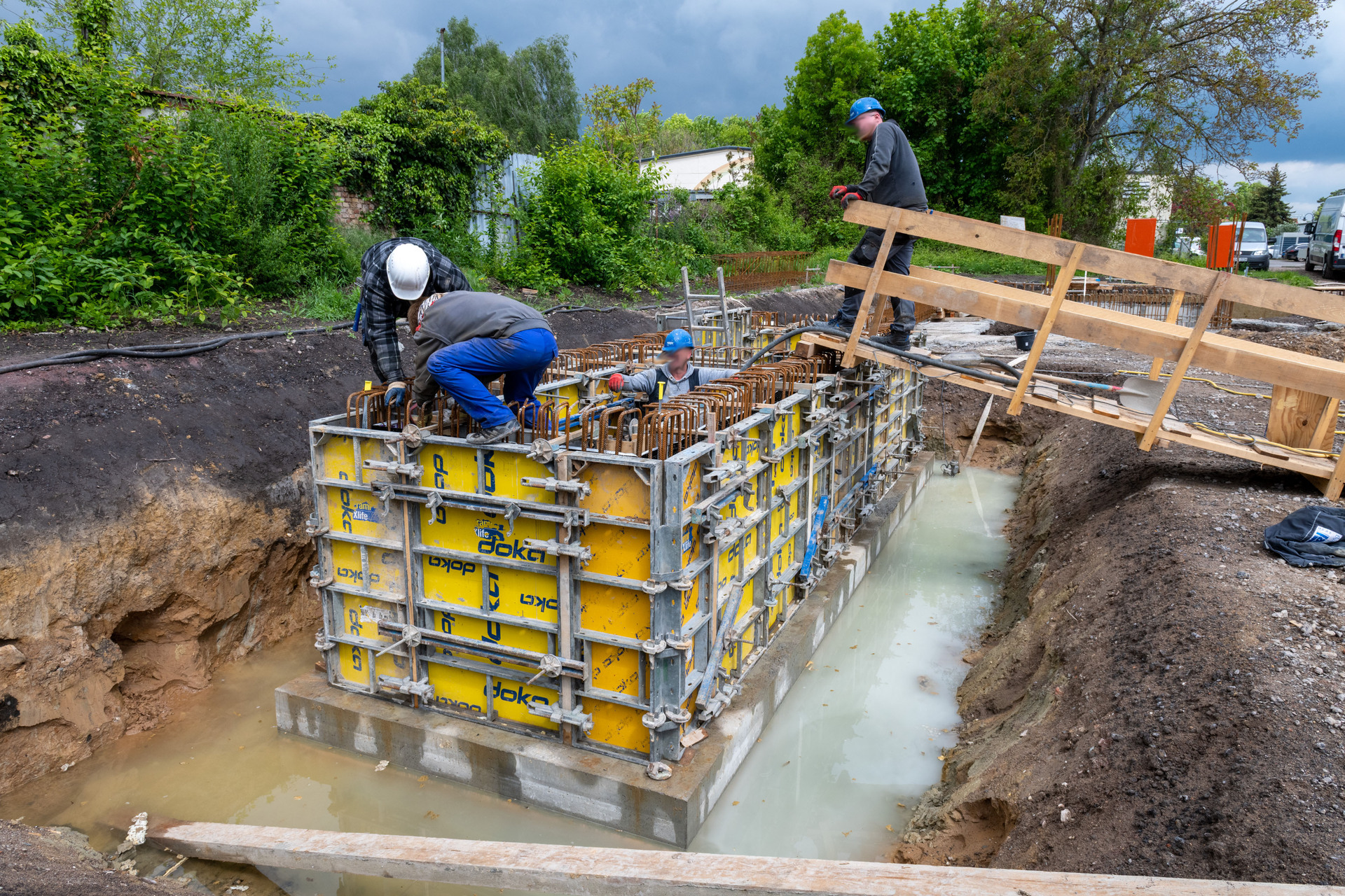 Drei Bauarbeiter montieren eine gelbe Schalung für eine Betonfundamentkonstruktion in einer ausgehobenen Baugrube mit Wasseransammlung, umgeben von Bewehrungsstahl.