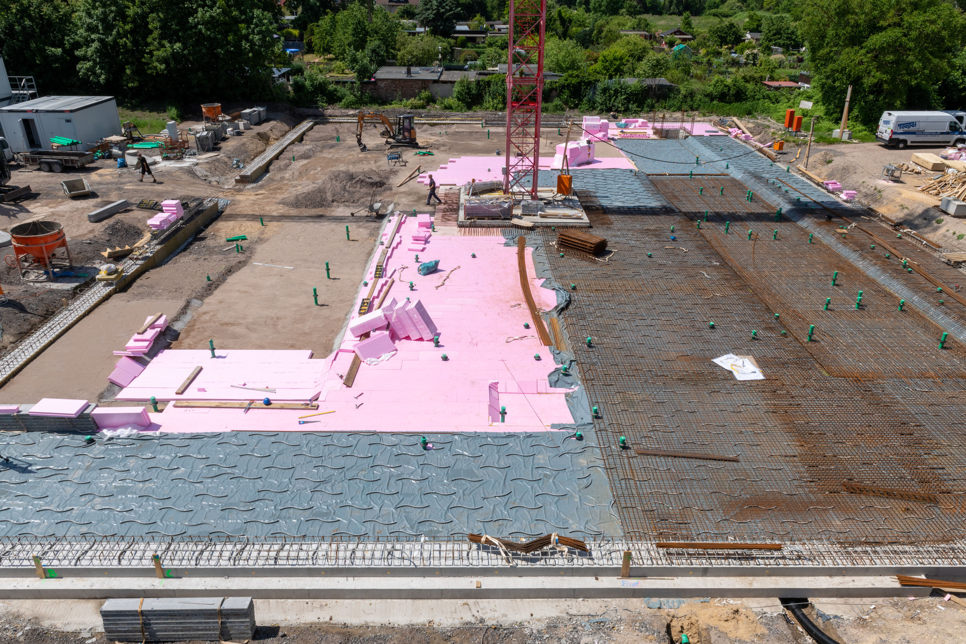 Betonfundament im Rohbau mit pinken Isolierplatten als Dämmung, Stahlbewehrung und vorbereiteten Schichten für Bodenbeläge. Zwei Bauarbeiter auf der Baustelle, umgeben von Baumaschinen und Materialien.