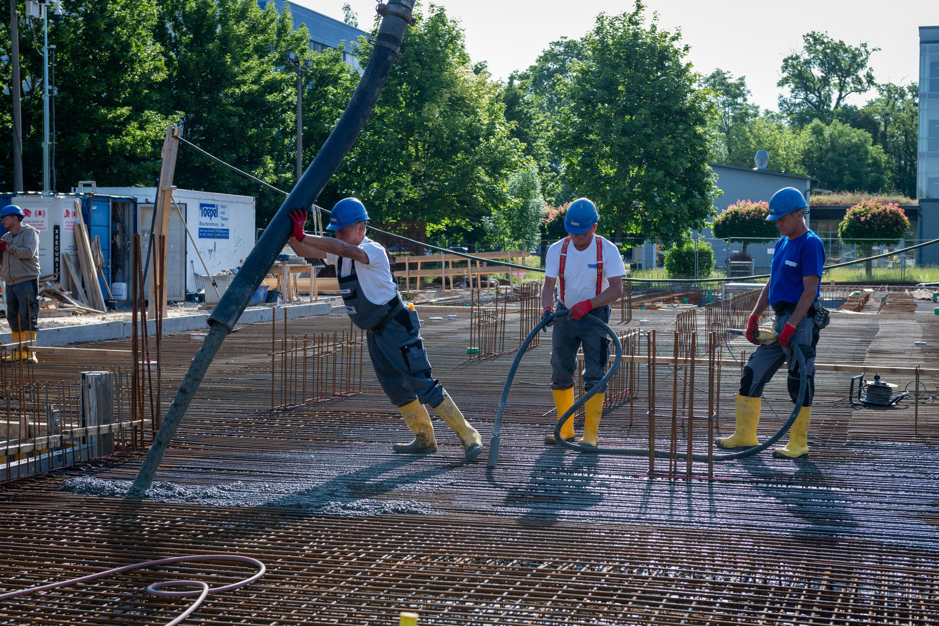Drei Bauarbeiter auf einer Baustelle, die gemeinsam eine lange Stahlstange in ein Betonfundament oder eine Schalung einführen, alle mit Schutzhelmen, Handschuhen und gelben Sicherheitsschuhen.