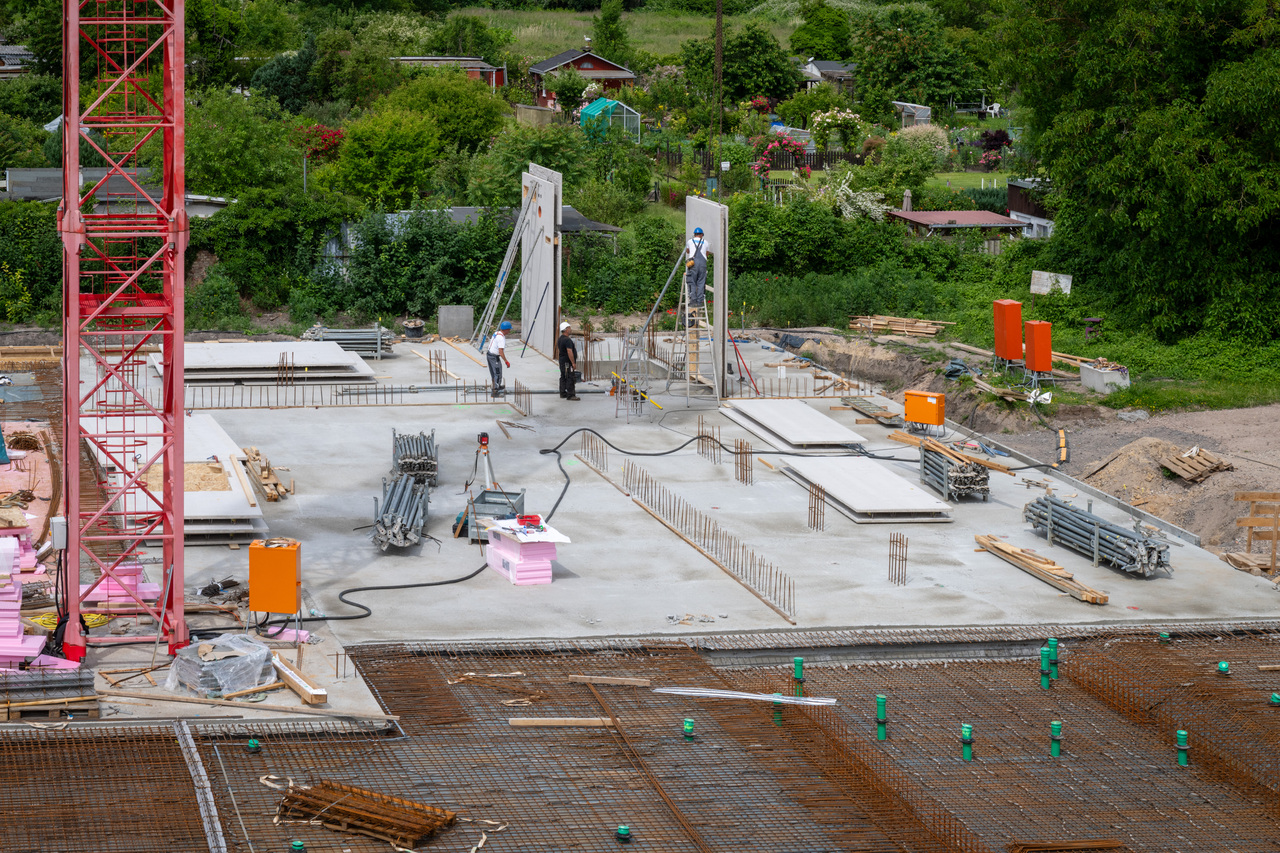 Auf einer Baustelle werden Betonfundamente gegossen. Drei Arbeiter positionieren Bewehrungseisen und montieren Schalungen, während im Hintergrund ein roter Baukran steht und Materialien wie Holz und Stahlrohre verteilt liegen.