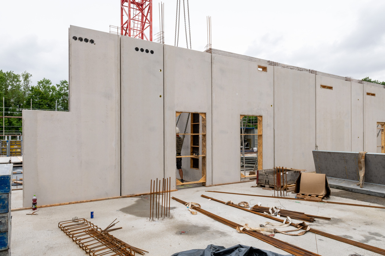 Rohbau eines Gebäudes mit Betonwänden, zwei offenen Fenster- und Türöffnungen, die mit Holz verkleidet sind. Vor der Wand liegen Bewehrungsstähle und Baustahlmatten auf dem Boden. Im Hintergrund ein roter Baukran.