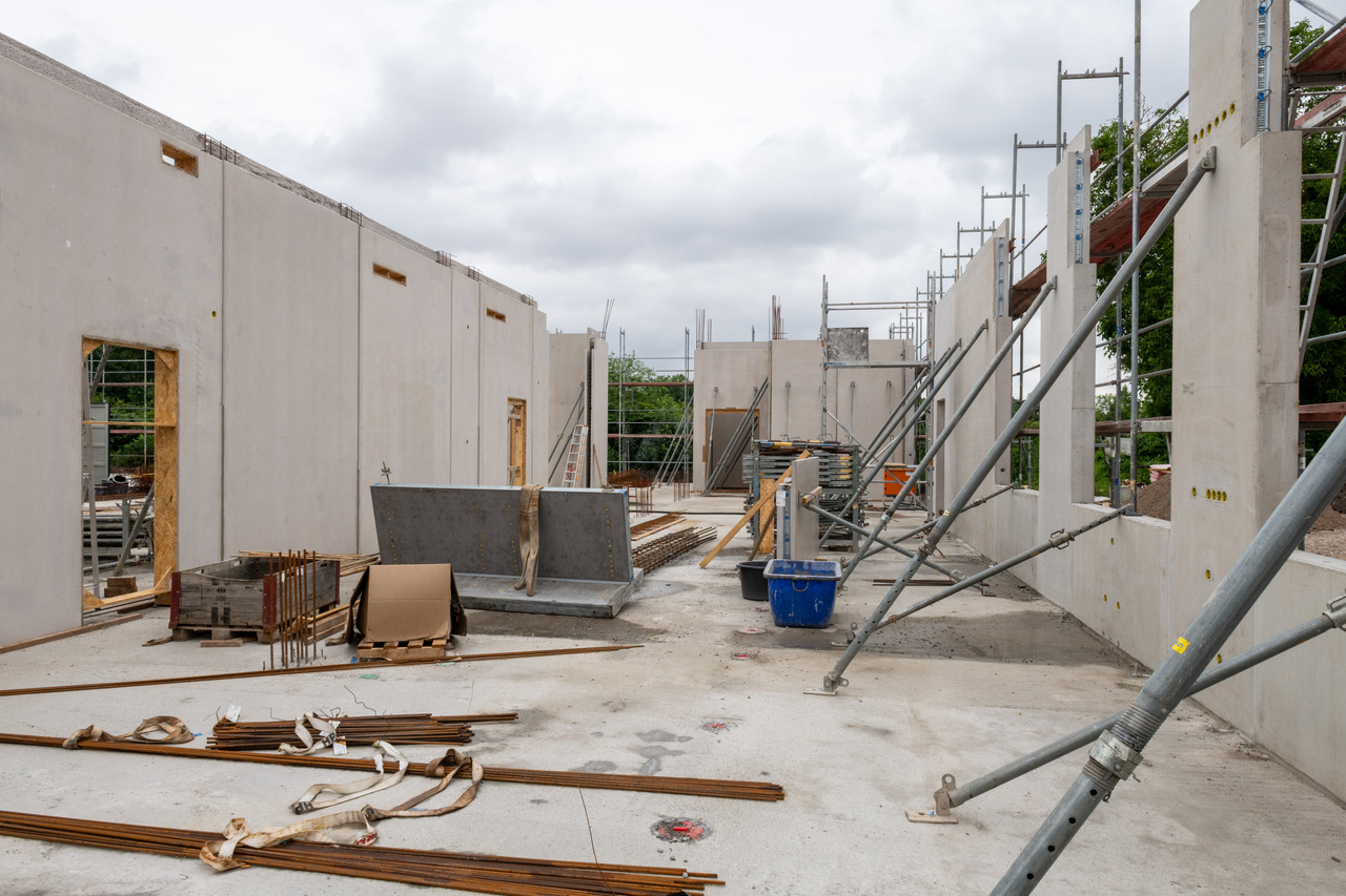 Baustelle mit Rohbau eines mehrstöckigen Gebäudes, teilweise verputzte Wände und offene Fensteröffnungen. Im Vordergrund liegen Baustahlmatten, Rohre und Werkzeug. Eine blaue Kiste und eine Leiter sind sichtbar, im Hintergrund Gerüste.
