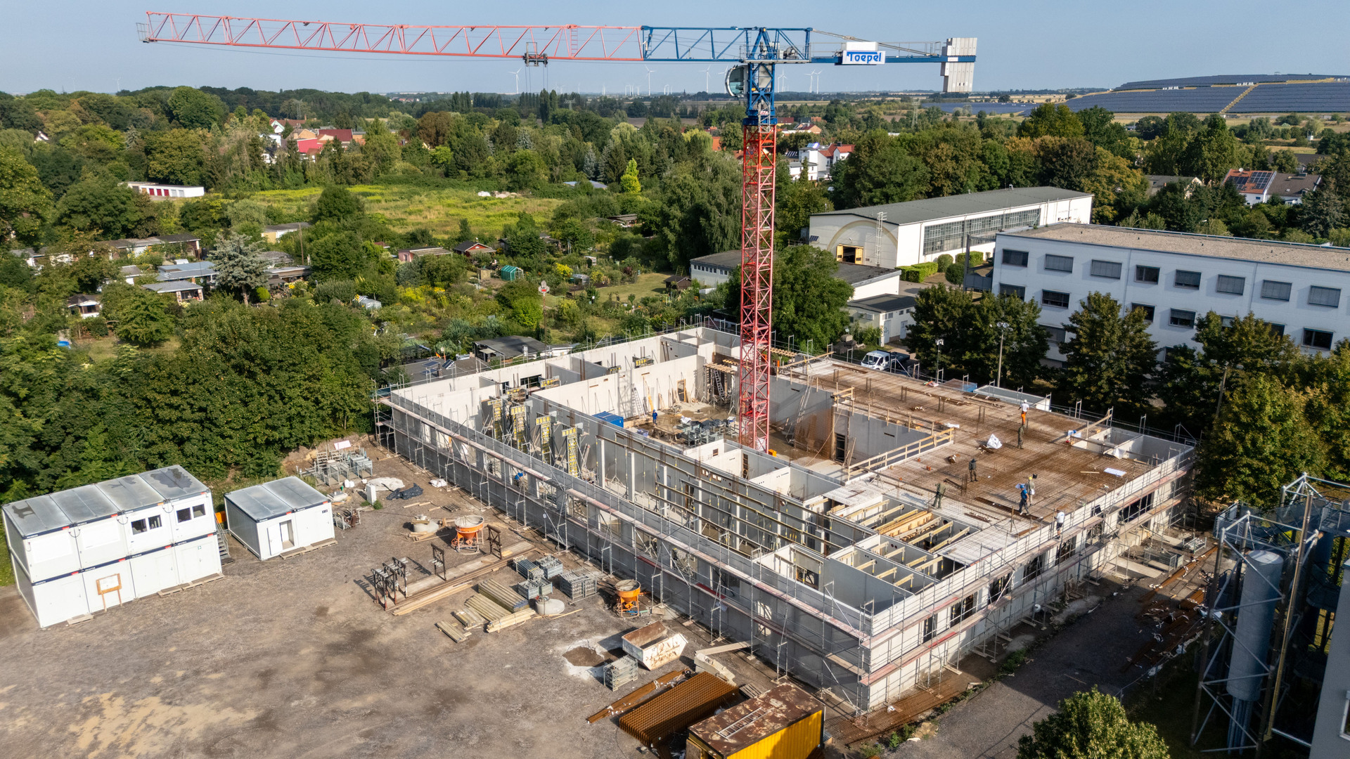 Baugebiet mit einem mehrstöckigen Rohbau im Rohzustand, umgeben von Gerüsten und Baustelleneinrichtungen. Ein großer Turmkran ist im Einsatz, daneben Container und Baumaschinen. Im Hintergrund grüne Wald- und Wiesenflächen sowie weitere Gebäude.