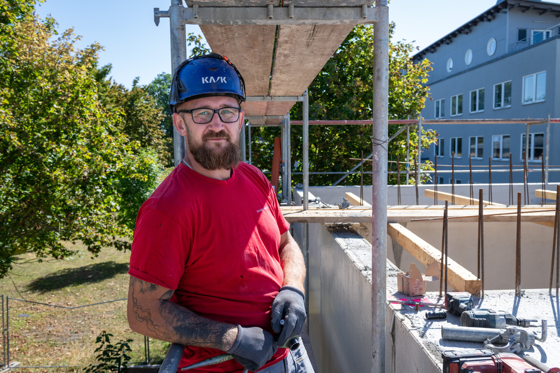 Ein Bauarbeiter mit Schutzhelm und roter Arbeitskleidung steht auf einer Baustelle. Er hält sich an einer Metallkonstruktion fest und lächelt zur Kamera. Im Hintergrund sind Gerüste, Baustoffe und ein Gebäude im Rohbau zu sehen.
