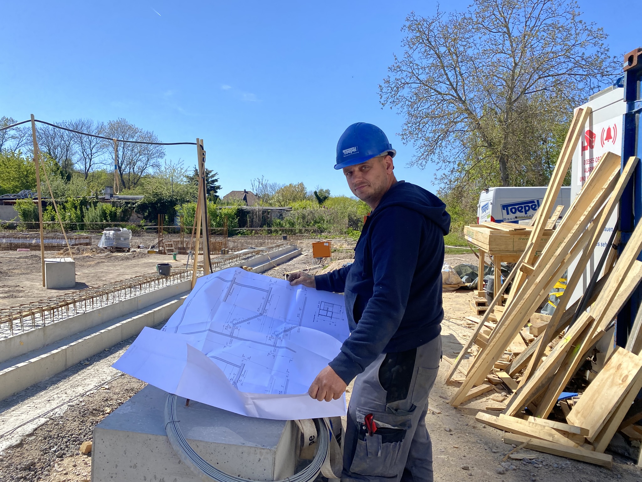 Ein Bauarbeiter in blauer Schutzhelm und Arbeitskleidung hält einen großen Bauplan und steht auf einer Baustelle mit sichtbaren Betonfundamenten und Holzgerüsten.