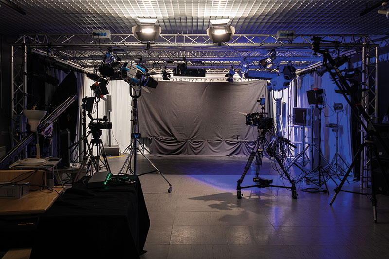 Großer leerer Studiobereich mit hellgrauer Rückwand und seitlichen Vorhängen. Mehrere professionelle Kamerastative mit montierten Kameras und Beleuchtungseinrichtungen sind symmetrisch aufgestellt, teilweise mit Scheinwerfern und Reflektoren. Boden aus glattem Material, links ein dunkler Tisch.