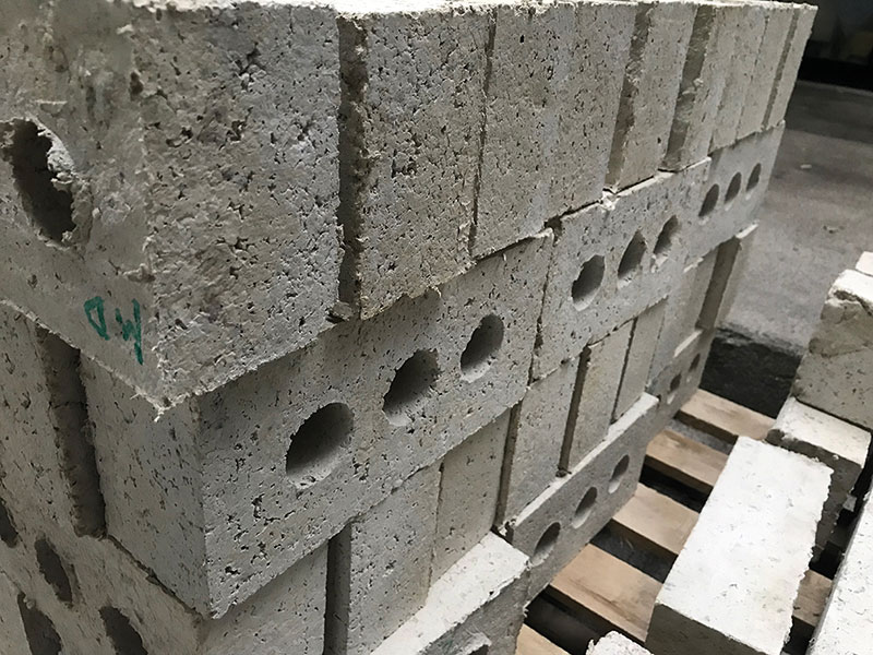 Graustichige Betonsteine mit mehreren runden Löchern in der Mitte, gestapelt in einer Reihe. Einige Steine zeigen Beschädigungen oder Absplitterungen an den Kanten. Die Steine liegen auf einer Holzpalette.