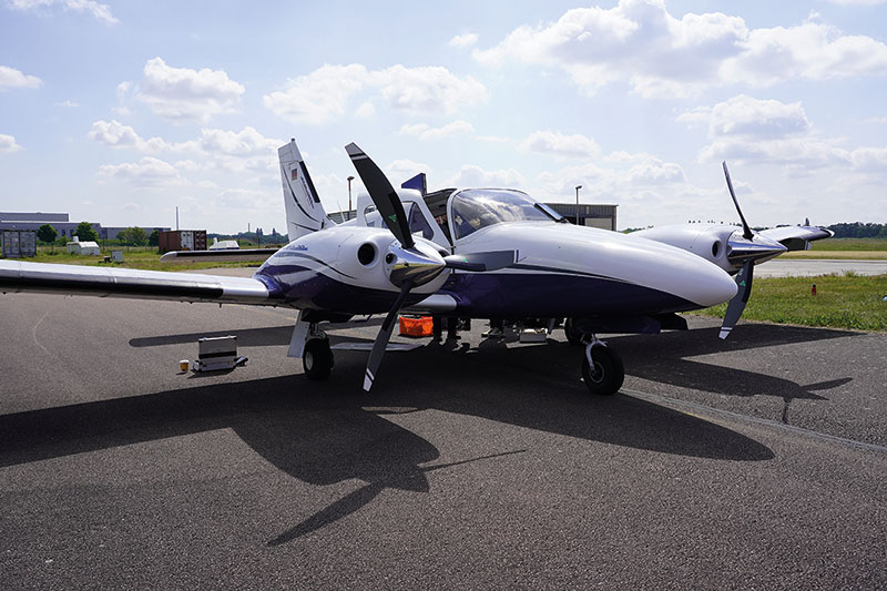 Kleines zweimotoriges Propellerflugzeug in weißer und grauer Farbe steht auf einem Rollfeld. Die Maschine hat ein T-Fahrwerk und ist mit zwei Propellern an den Tragflächen ausgestattet.