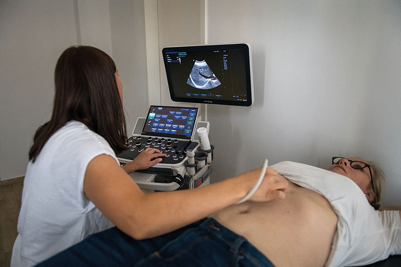 Eine Ärztin führt eine Ultraschalluntersuchung am Oberbauch einer liegenden Patientin durch, während auf einem Bildschirm eine Echtzeit-Echobilddarstellung zu sehen ist.