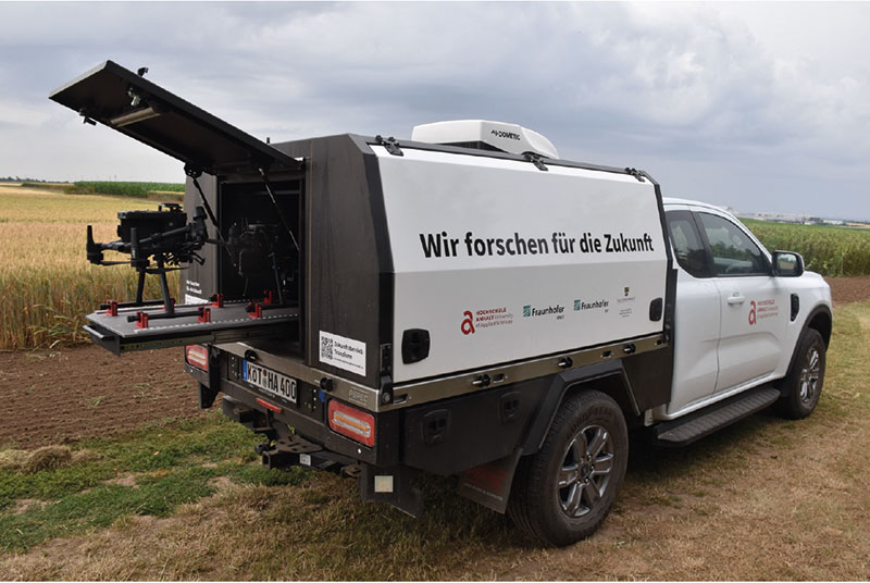Ein weißer Forschungspickup mit aufgebocktem Heckteil, ausgestattet mit technischer Messausrüstung. Auf der Ladefläche steht in Großbuchstaben Wir forschen für die Zukunft. Fahrzeug steht auf einem Feld.