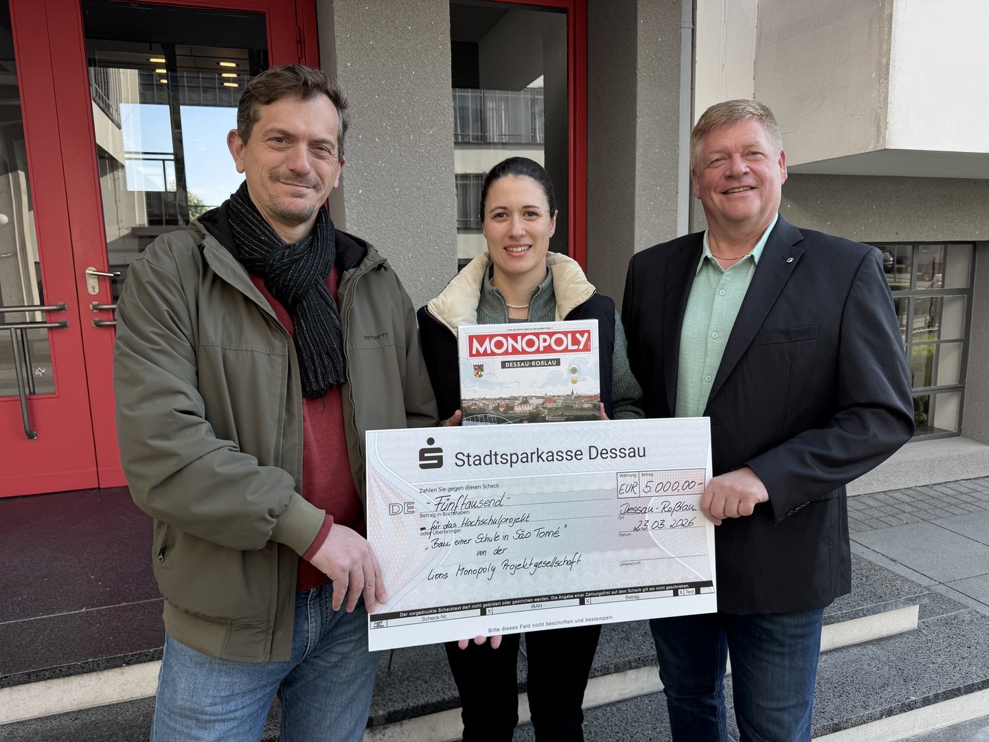 Drei Personen vor einer Stadtsparkasse Dessau: Ein Mann und eine Frau in der Mitte halten ein Schecküberreichungsdokument über 5.000 Euro für das Projekt „Monopoly“ mit Spielsteinen. Der Mann rechts trägt Anzug und Krawatte.