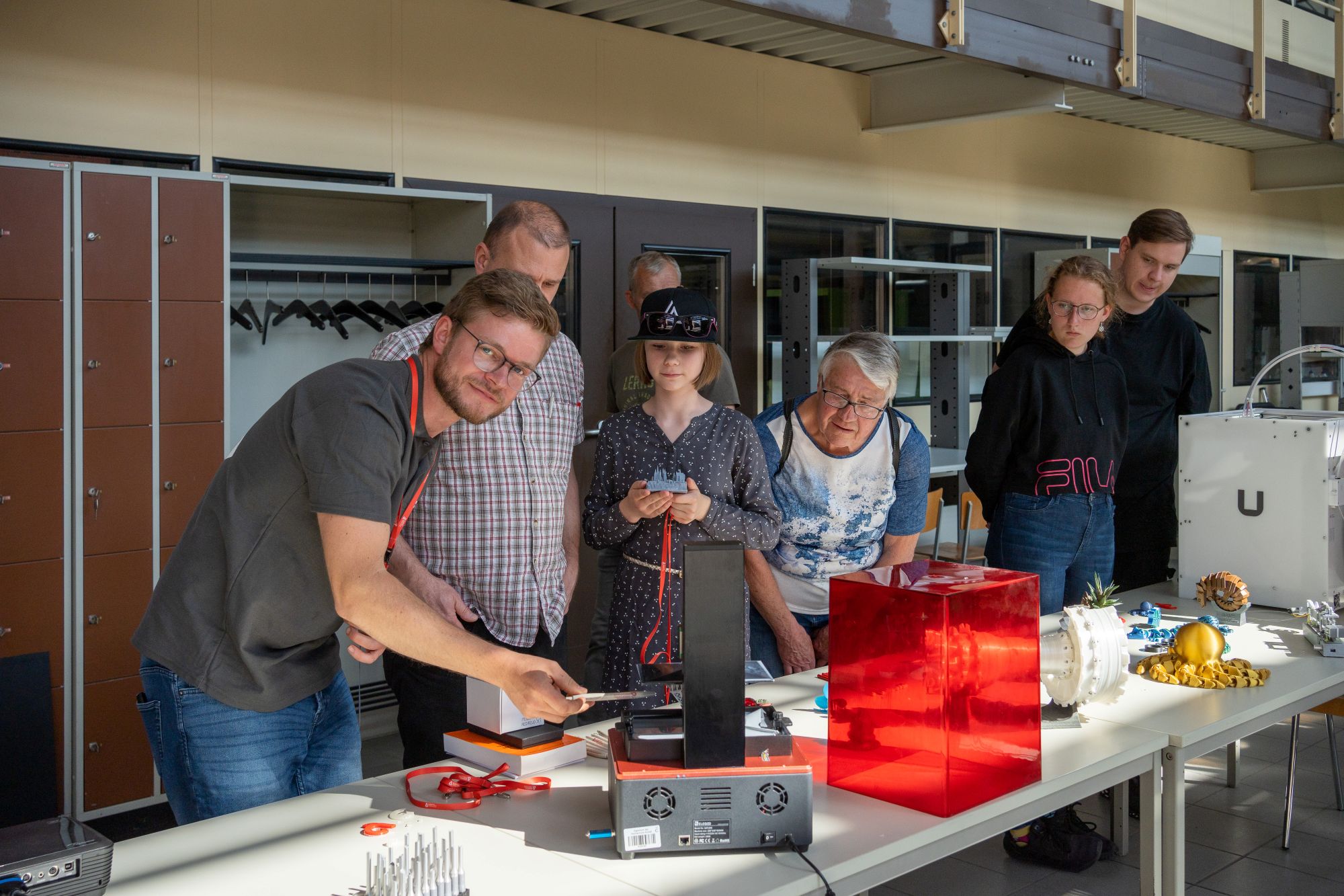 Eine Gruppe von sechs Personen beobachtet und unterstützt einen Mann, der einen 3D-Drucker bedient, der gerade ein Objekt aus schwarzem Filament herstellt. Auf dem Tisch liegen weitere gedruckte Modelle, darunter ein roter Behälter.