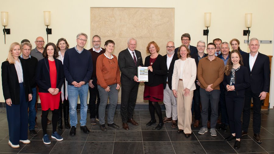 Gruppe von 19 Personen, darunter Männer und Frauen, steht lächelnd für ein Gruppenfoto. Ein Mann in der Mitte hält ein Buch oder eine Urkunde. Alle tragen formelle oder geschäftliche Kleidung.