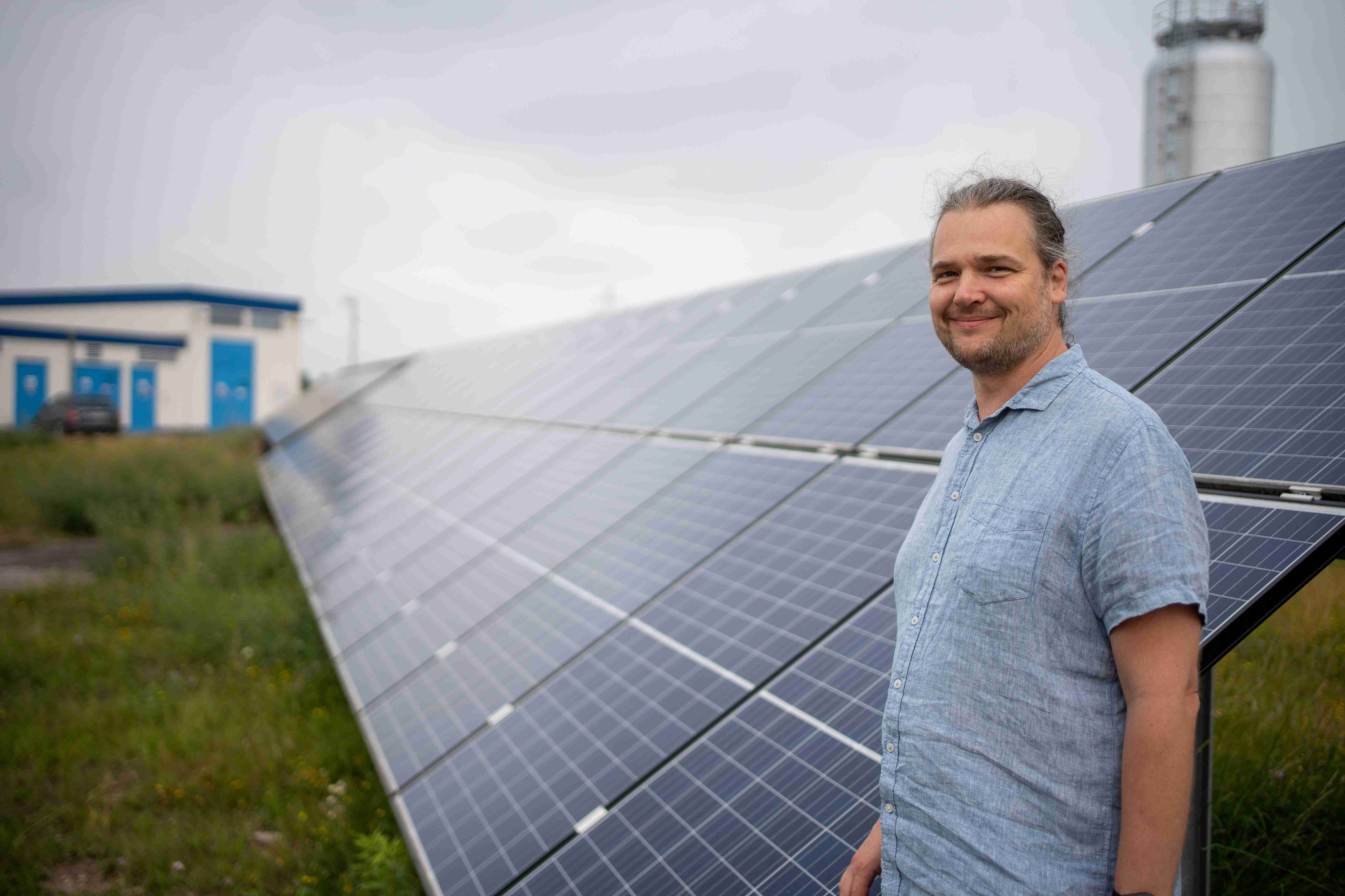 Ein lächelnder Mann steht vor einer großen Solaranlage mit vielen nebeneinander aufgestellten schwarzen Photovoltaik-Paneelen auf einem Feld.