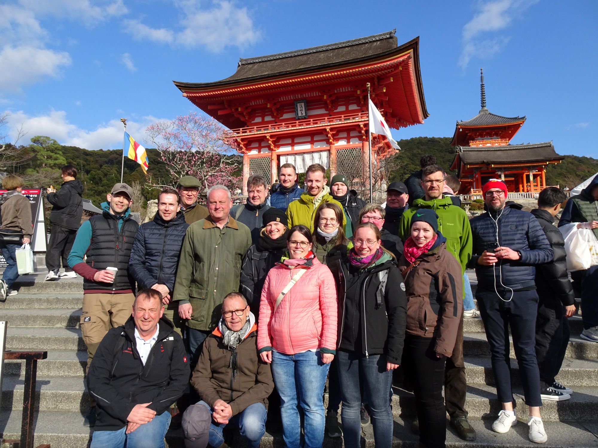 Gruppe von 16 Personen, darunter Männer und Frauen, posiert lächelnd vor dem berühmten roten Tor des Kiyomizu-dera in Kyoto, Japan. Im Hintergrund sind traditionelle japanische Architektur und Bäume mit blühenden Zweigen zu sehen.