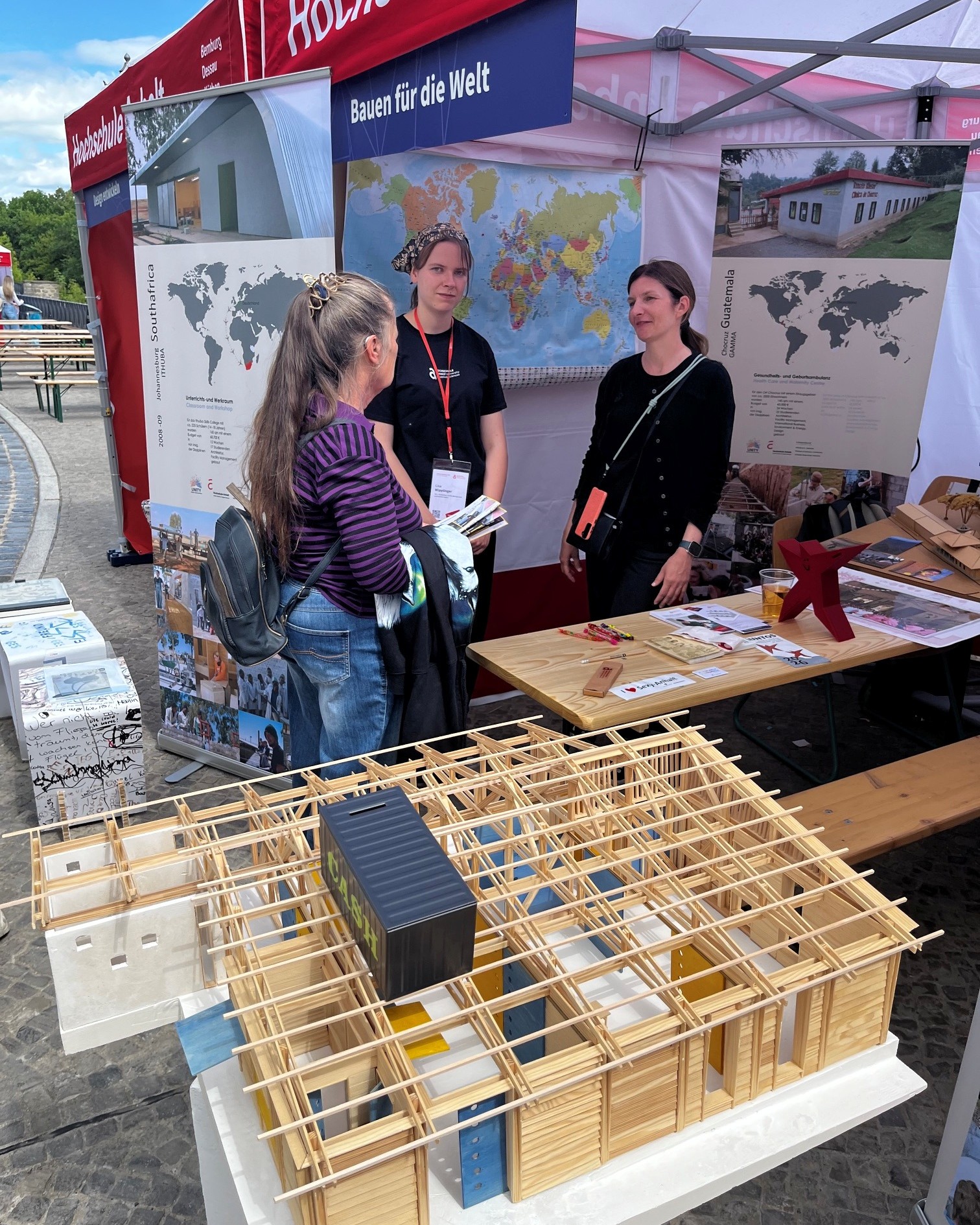 Drei Personen stehen an einem Infostand mit dem Thema „Bauen für die Welt“. Vor ihnen liegt ein großes Holzmodell eines Gebäudes mit verschiedenen farbigen Elementen, das nachhaltiges Bauen oder eine Bauweise für Entwicklungsprojekte darstellt.