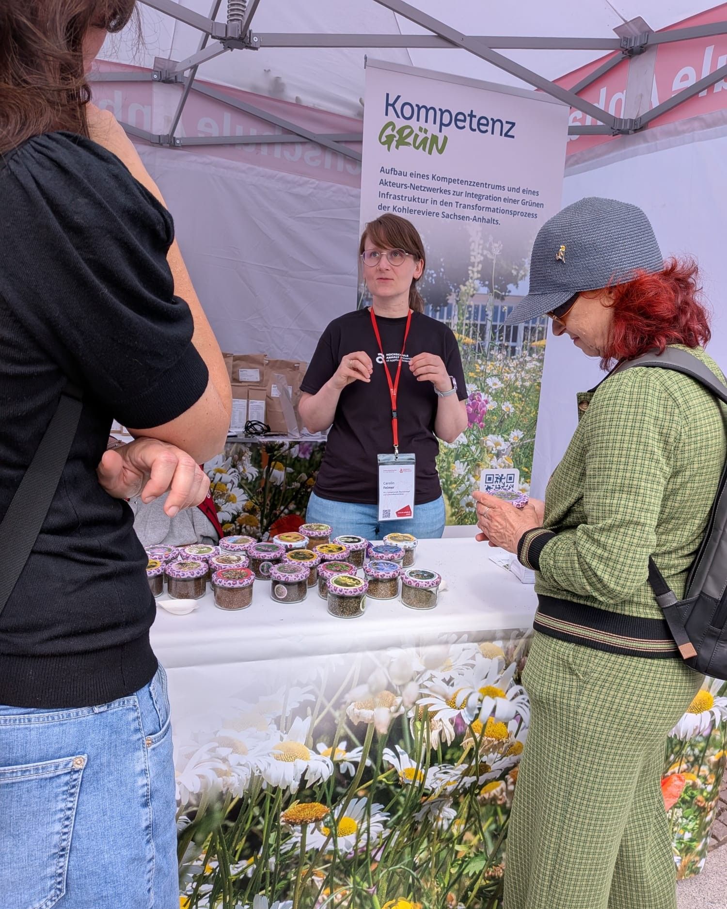 Zwei Personen unterhalten sich an einem Infostand mit dem Titel „KompetenzGrün“. Auf dem Tisch stehen mehrere Gläser mit eingekochten Produkten, vermutlich Marmelade oder Konfitüre, umgeben von gelben und weißen Blumen. Eine dritte Person steht hinter dem Tisch und informiert.