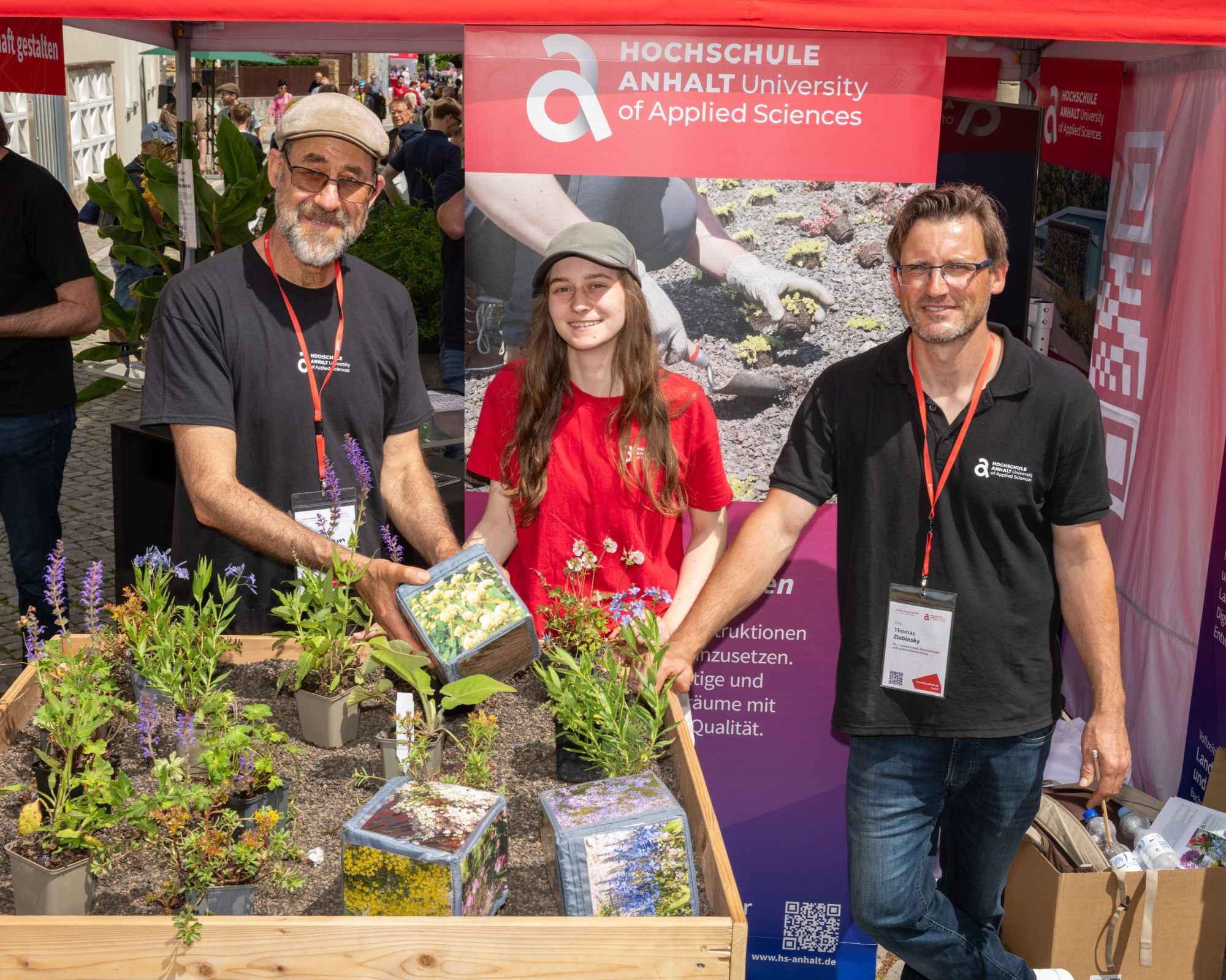 Drei Personen stehen vor einem Stand der Hochschule Anhalt – University of Applied Sciences. Sie halten und präsentieren verschiedene Pflanzen in Töpfen und Verpackungen, lächelnd und in lockerer Atmosphäre.