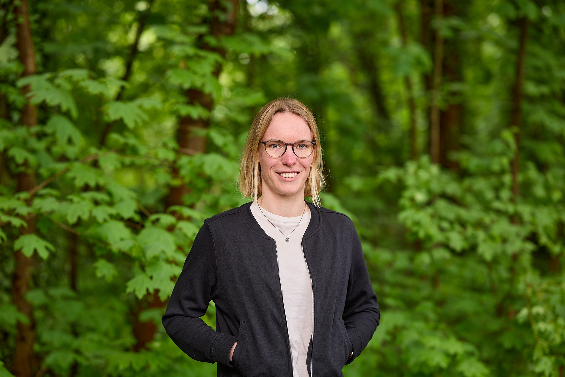 Eine lächelnde Person mit hellen Haaren und Brille steht vor einem grünen Waldhintergrund, trägt einen dunklen Blazer über einem weißen Shirt und hat die Hände in den Taschen.
