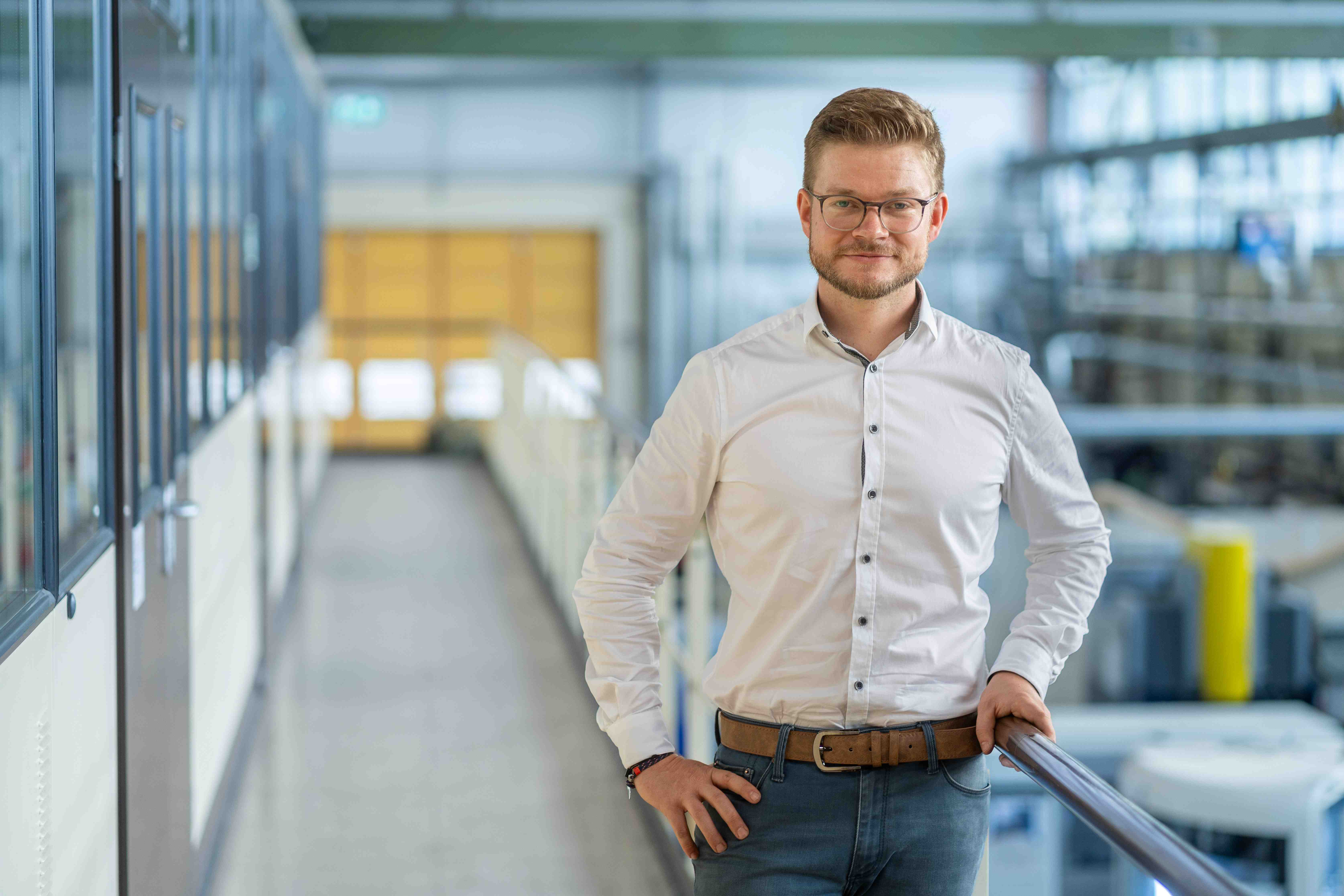 Foto zeigt Mann mit Brille und weißem Hemd am Geländer eines industrielle wirkenden Labors an der Hochschule Anhalt, in dem an 3D-Druck für Ersatzbauteile aus Metall geforscht wird
