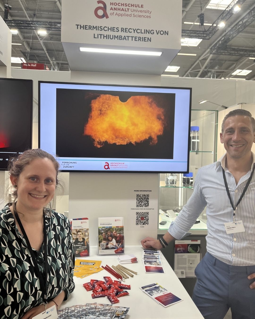 Zwei Personen stehen lächelnd an einem Infostand der Hochschule Anhalt zum Thema Lithium-Batterien recyceln. Im Hintergrund ist eine Explosionsdarstellung auf einem Bildschirm zu sehen, auf dem Tisch liegen Informationsmaterialien und Batterien.