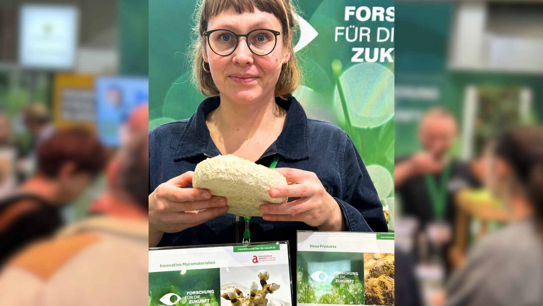 Foto mit Person, die weiß-gelbes, schwammähnliches Pilzmyzel als Verpackung in der Hand hält, zeigt Anna Gronemeyer von der Hochschule Anhalt. Im Hintergrund Messesituation von der Grünen Woche. 