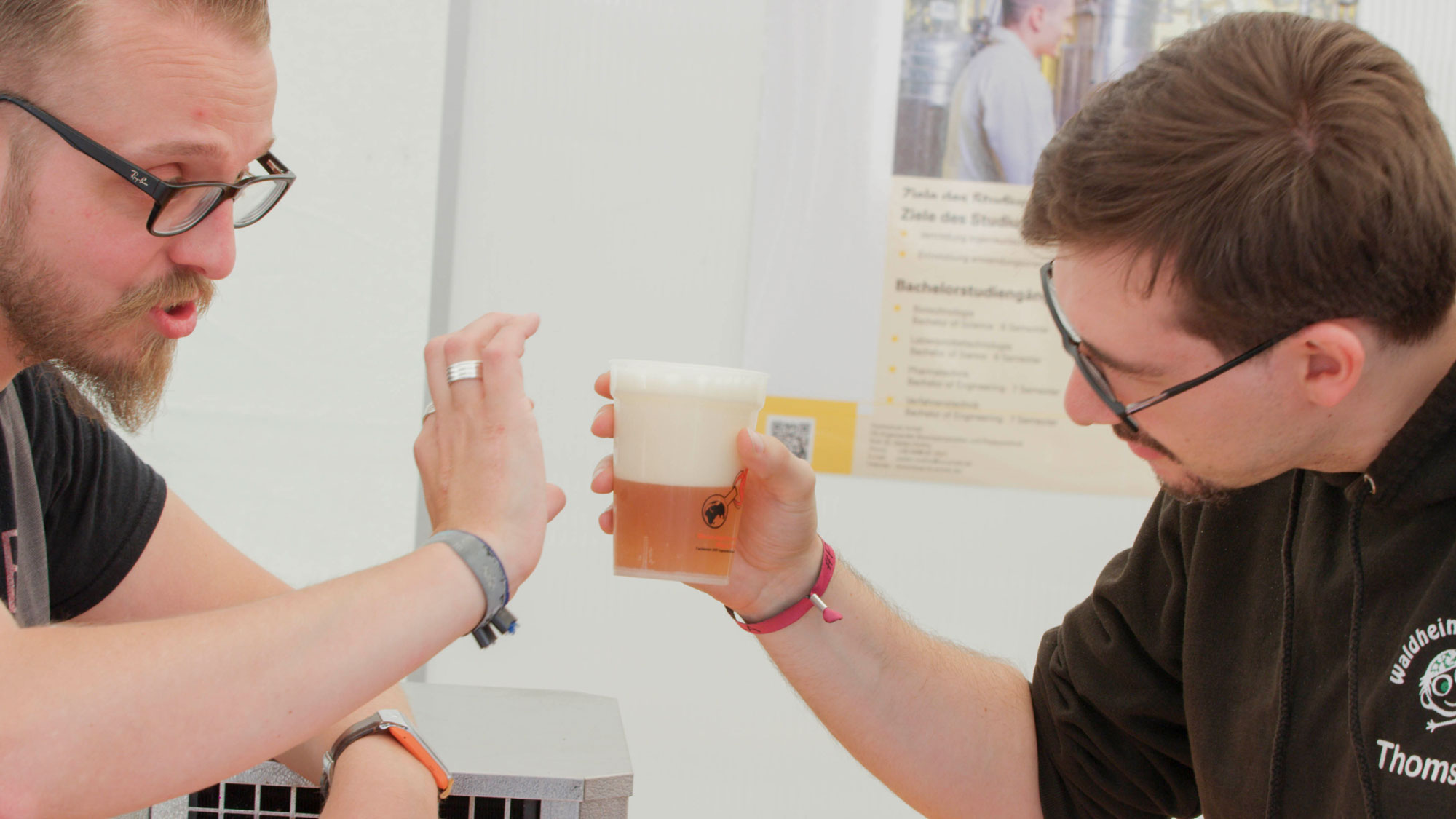 Zwei Männer stoßen mit Gläsern an, eines davon enthält ein helles, schäumendes Getränk, vermutlich Bier. Sie wirken freundlich und sind in einer Innenraumumgebung mit Informationsplakaten im Hintergrund.