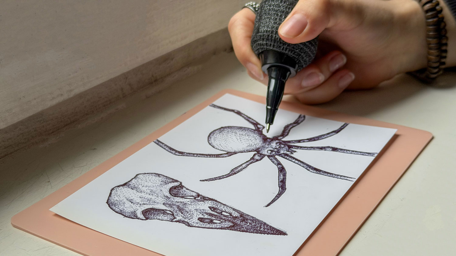 Eine Hand zeichnet mit einem Fineliner eine Spinne mit acht langen Beinen und einem detaillierten Körper auf ein Blatt Papier, daneben eine Skizze eines Hundeschädels.