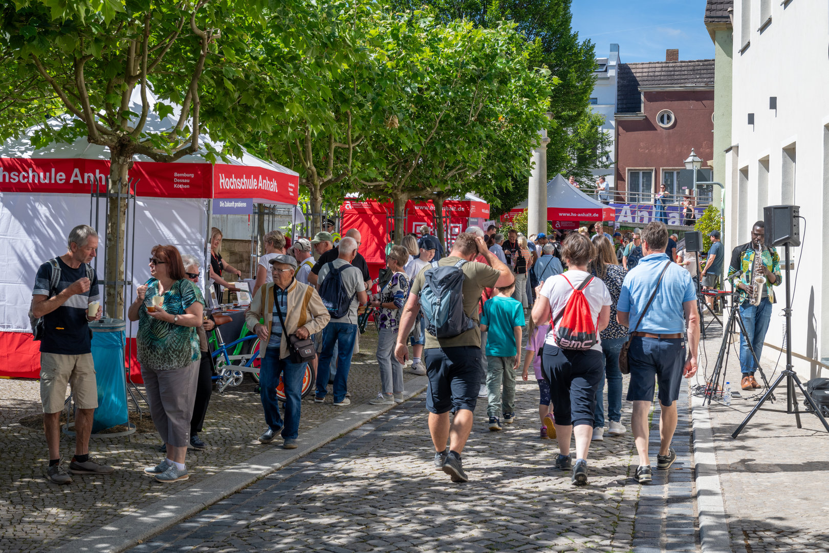 Belebte Informationsmesse mit vielen Besuchern, die sich zwischen roten Infoständen der Hochschule Anhalt bewegen, darunter auch ein Musikstand mit Live-Auftritt.