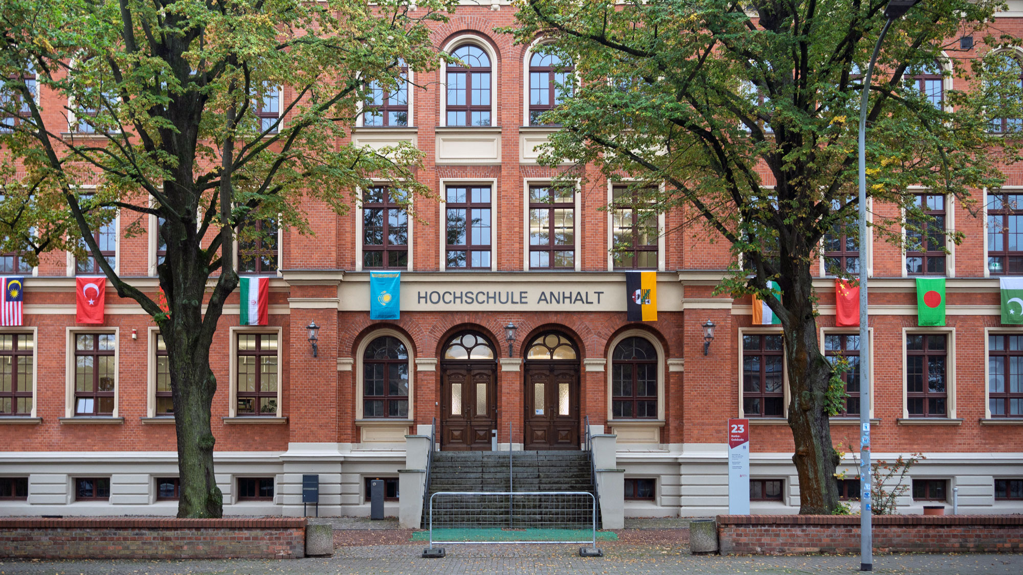 Rotes Backsteingebäude mit zentralem Haupteingang und Treppe, beschriftet als „Hochschule Anhalt“. Mehrere Flaggen internationaler Partnerhochschulen hängen an der Fassade.