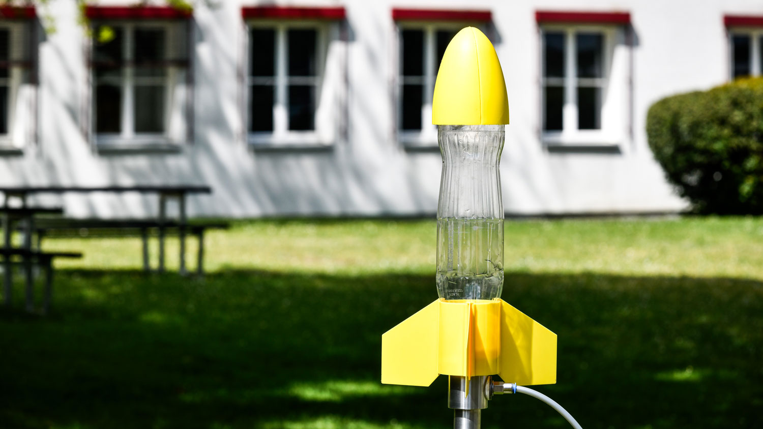 Gelbfarbene Modellrakete mit durchsichtigem Rumpf, die auf einem Ständer vor einem Gebäude mit Fenstern und Rasen steht.
