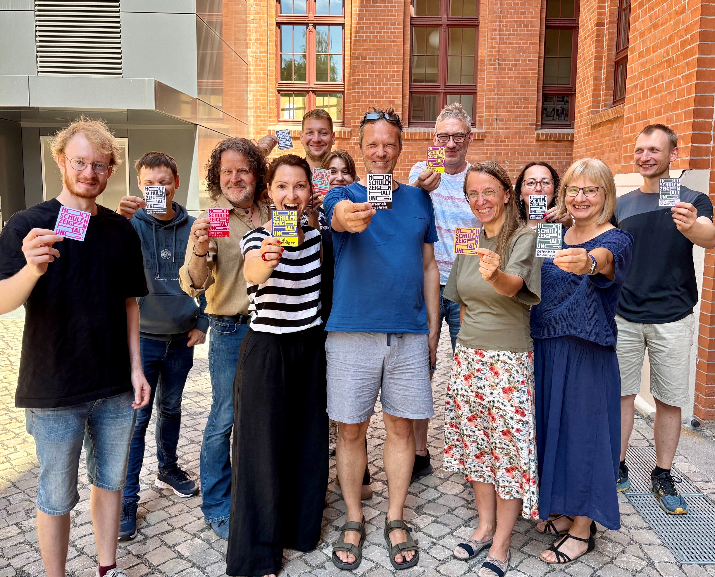 Gruppe von 14 Personen, die vor einem Gebäude stehen und jeweils bunte, kleine Karten oder Sticker in der Hand halten, lächelnd für ein Gruppenfoto.
