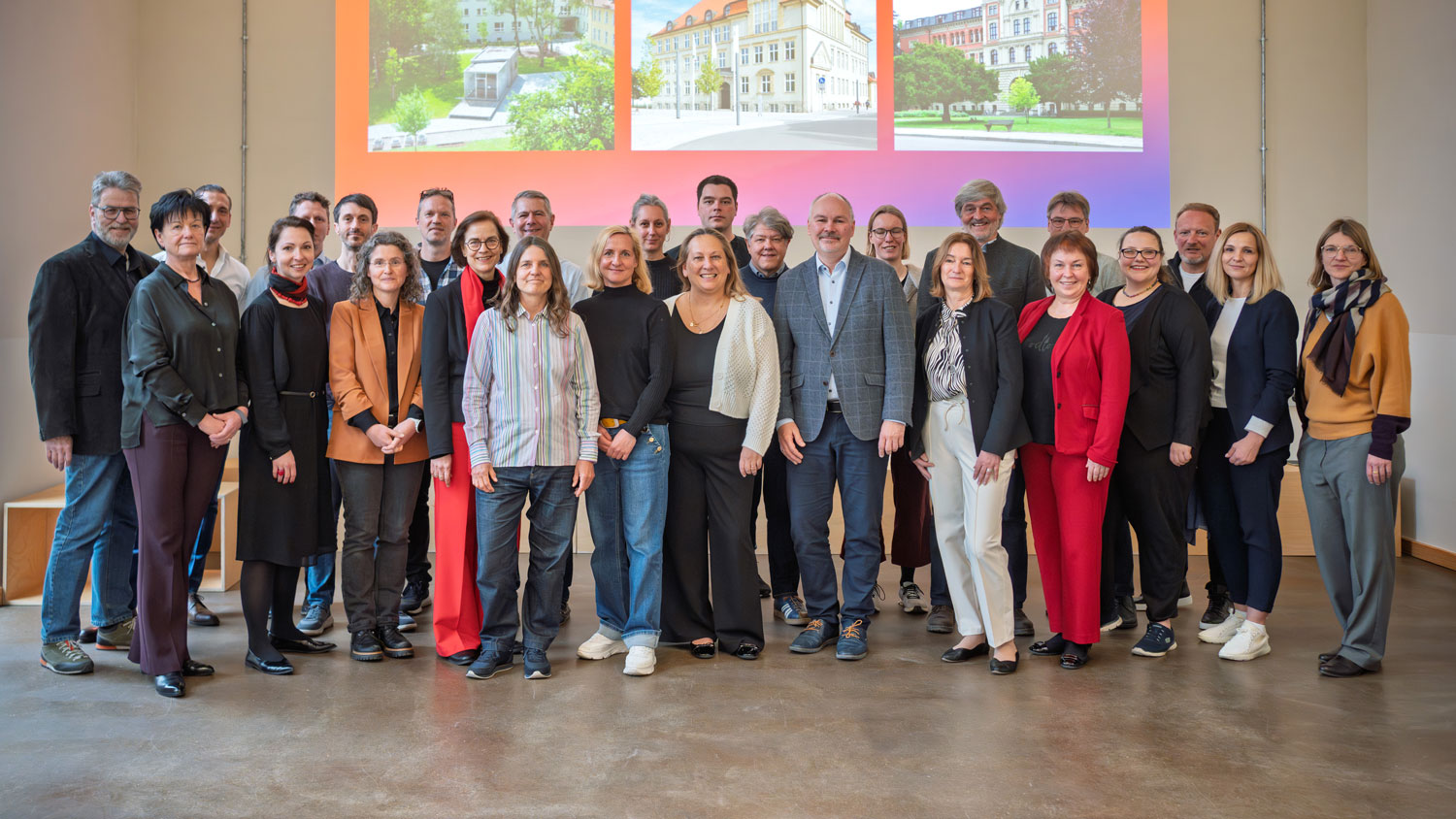 Gruppenfoto mit 20 Personen, die vor einem Hintergrund mit drei Bildern eines Gebäudes und eines grünen Platzes stehen. Die Gruppe besteht aus Männern und Frauen unterschiedlichen Alters, die lächeln und sich für ein gemeinsames Foto aufgestellt haben.