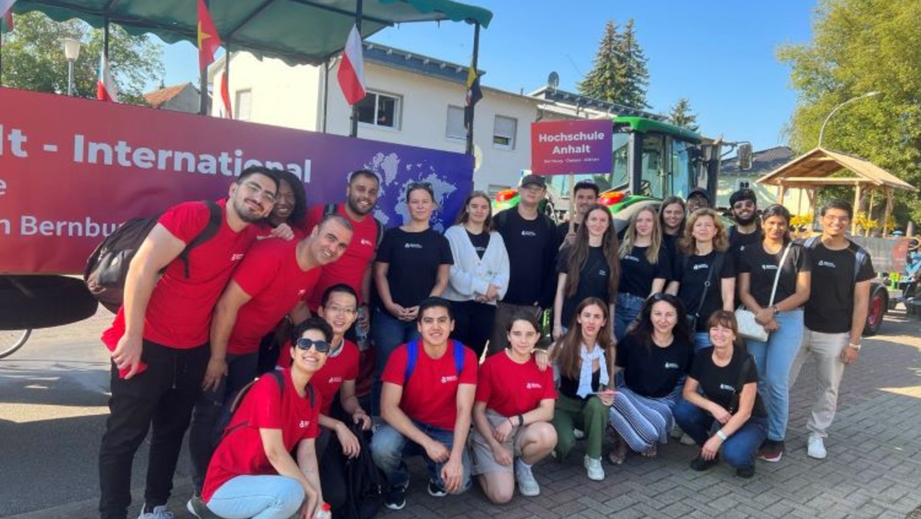 Eine Gruppe von Menschen steht vor einem Bus mit der Aufschrift "International" und "Bernburg". Die Personen tragen rote und schwarze T-Shirts und lächeln in die Kamera. Sie scheinen Teil einer Veranstaltung oder eines Projekts zu sein. Die Atmosphäre wirkt freundlich und gesellig.