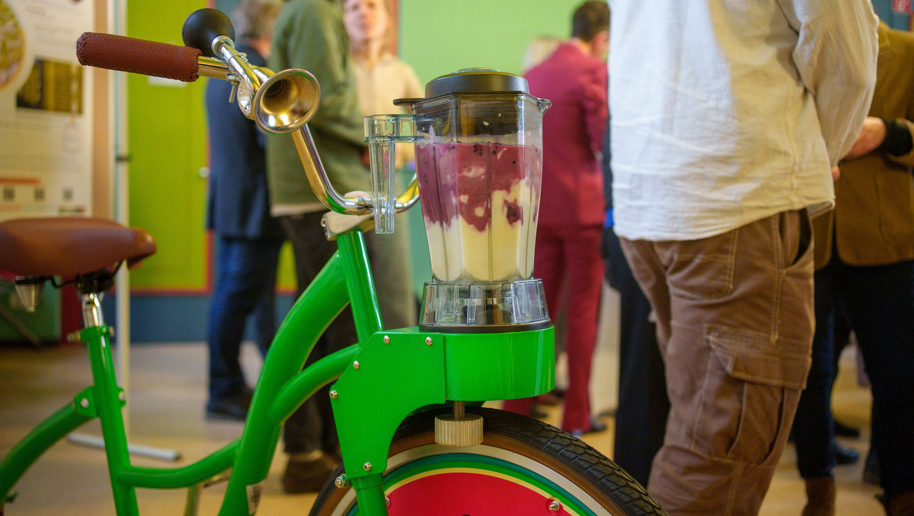 Grünes Fahrrad mit anmontiertem Mixer, in dem eine rote Fruchtmischung zu Smoothie verarbeitet wird.