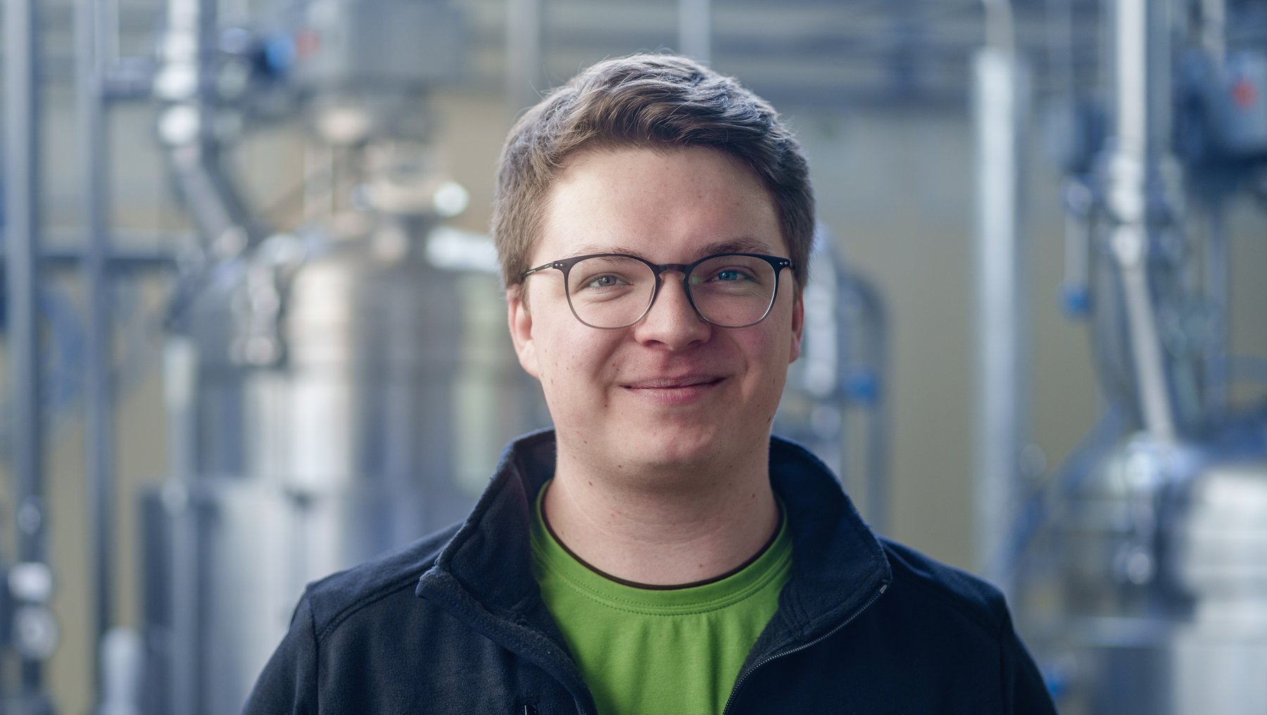 Ein lächelnder Mann mit Brille steht vor einer Industrieanlage. Er trägt eine schwarze Jacke und ein grünes T-Shirt. Der Hintergrund zeigt technische Geräte und Rohrleitungen, was auf eine Produktions- oder Laborumgebung hindeutet.