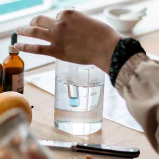 Eine Person hält eine Pipette über ein durchsichtiges Messbecherglas mit blauer Flüssigkeit, während zwei braune Tropfflaschen danebenstehen. Im Hintergrund liegt eine Orange.
