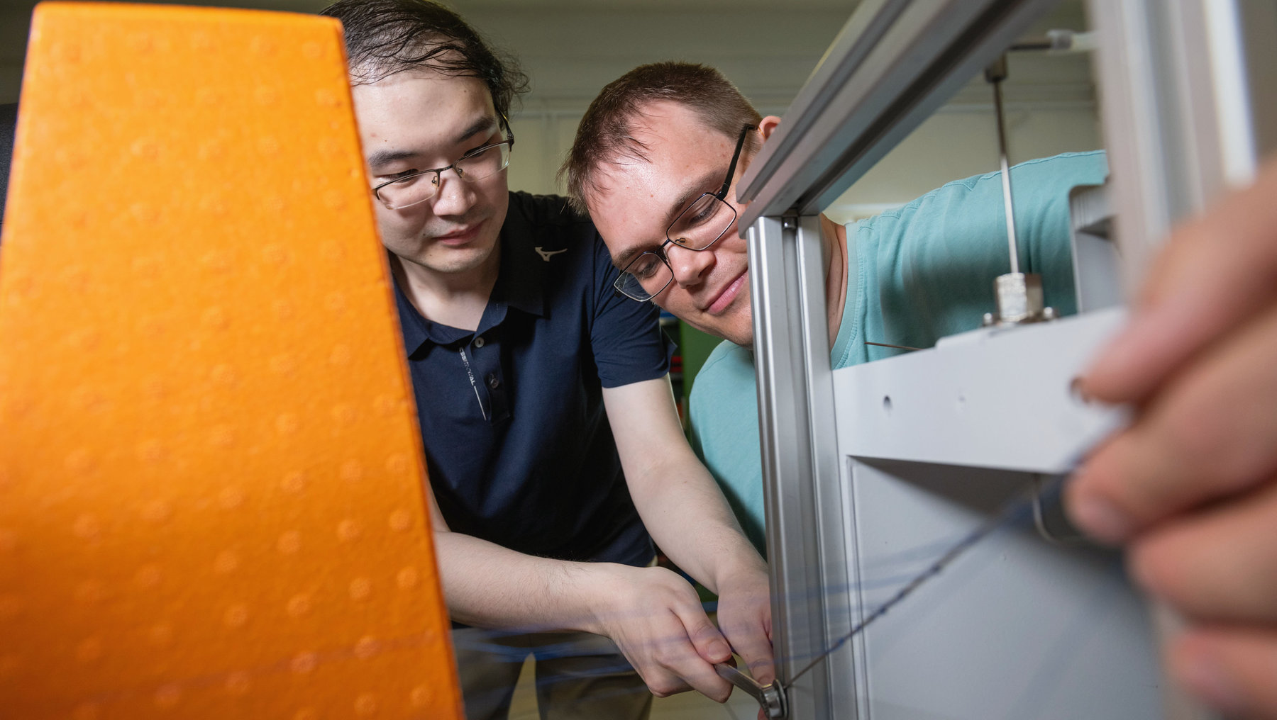 Zwei Personen arbeiten konzentriert an einem großen, orangefarbenen Gerät, das wie ein medizinisches oder wissenschaftliches Gerät aussieht. Sie halten gemeinsam eine Vorrichtung oder ein Werkzeug und scheinen etwas einzustellen oder zu justieren. Im Hintergrund ist eine helle Arbeitsumgebung mit Labor- oder Klinikcharakter zu erkennen.