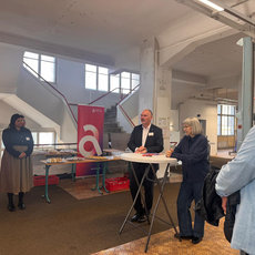 Fünf Personen stehen in einem hellen Industriegebäude mit großen Fenstern und Betonelementen. Ein Mann in Anzug spricht hinter einem Tisch mit Mikrofon, während die anderen zuhören – eine Frau mit grauen Haaren steht ihm direkt gegenüber. Im Hintergrund hängt ein rotes Banner mit weißem „a“.