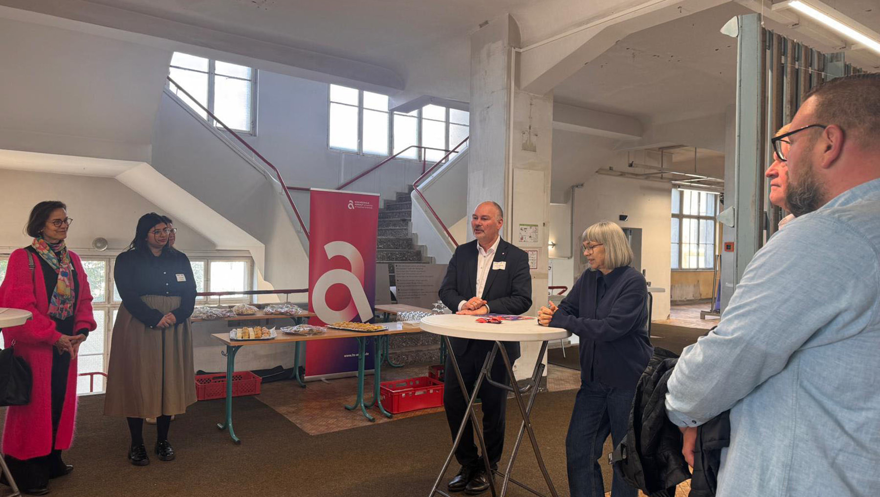 Fünf Personen stehen in einem hellen Industriegebäude mit großen Fenstern und Betonelementen. Ein Mann in Anzug spricht hinter einem Tisch mit Mikrofon, während die anderen zuhören – eine Frau mit grauen Haaren steht ihm direkt gegenüber. Im Hintergrund hängt ein rotes Banner mit weißem „a“.