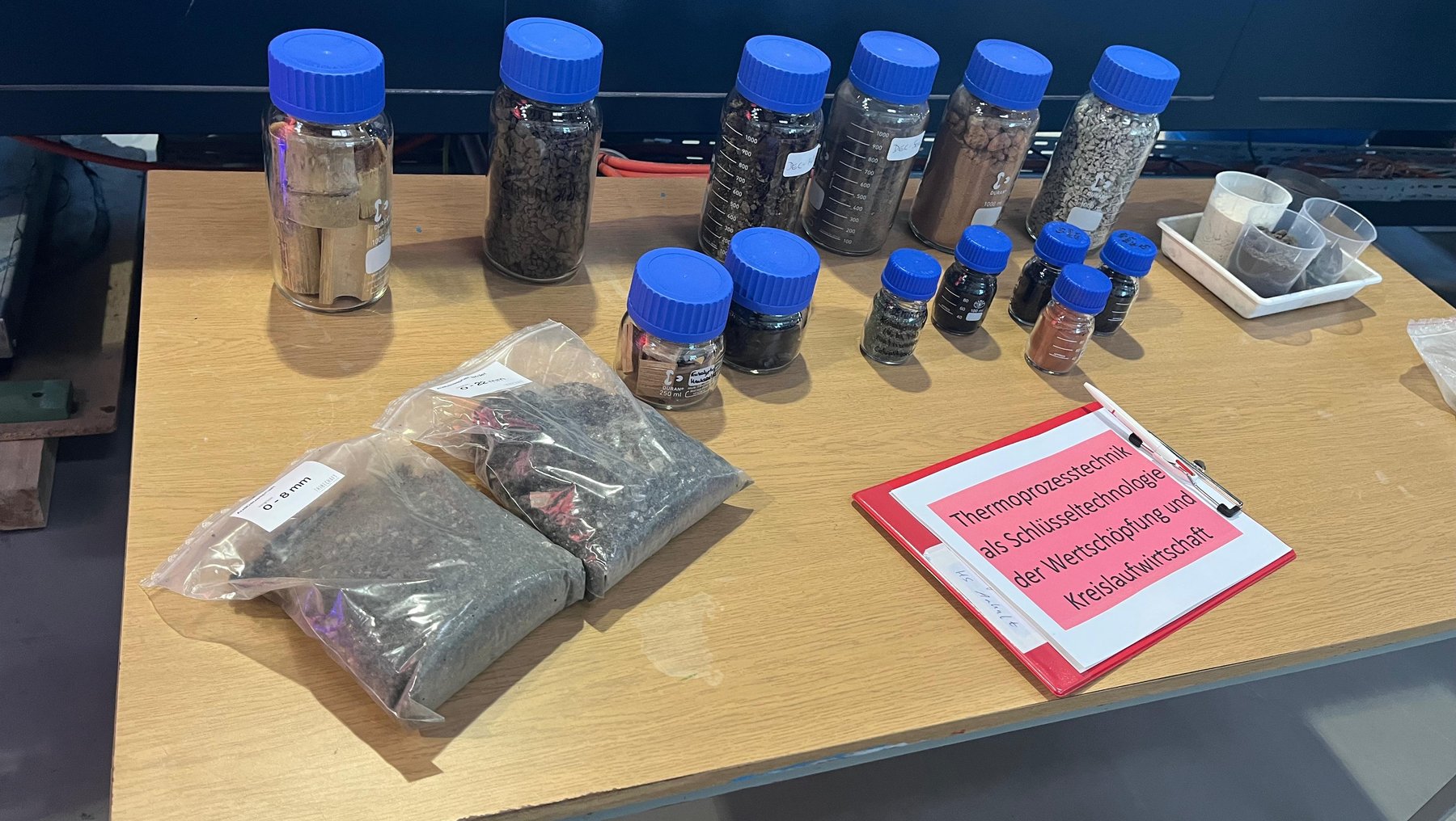 Auf einem Tisch stehen mehrere Gläser mit blauen Deckeln, gefüllt mit verschiedenen Samen oder kleinen Körnern. Daneben liegen verpackte Samenbeutel sowie ein rotes Schild mit der Aufschrift „Pflanzensamen zum Selbstbestäuben für Versuchsanbau“.