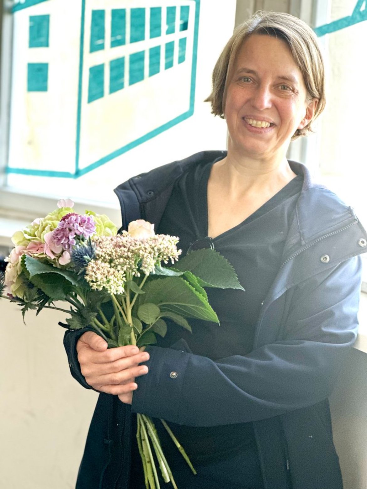 Dörte Meyer hält einen Strauß mit bunten Blumen, darunter weiße, lila und rosa Blüten sowie grüne Blätter, in den Händen. Sie trägt eine dunkle Jacke.