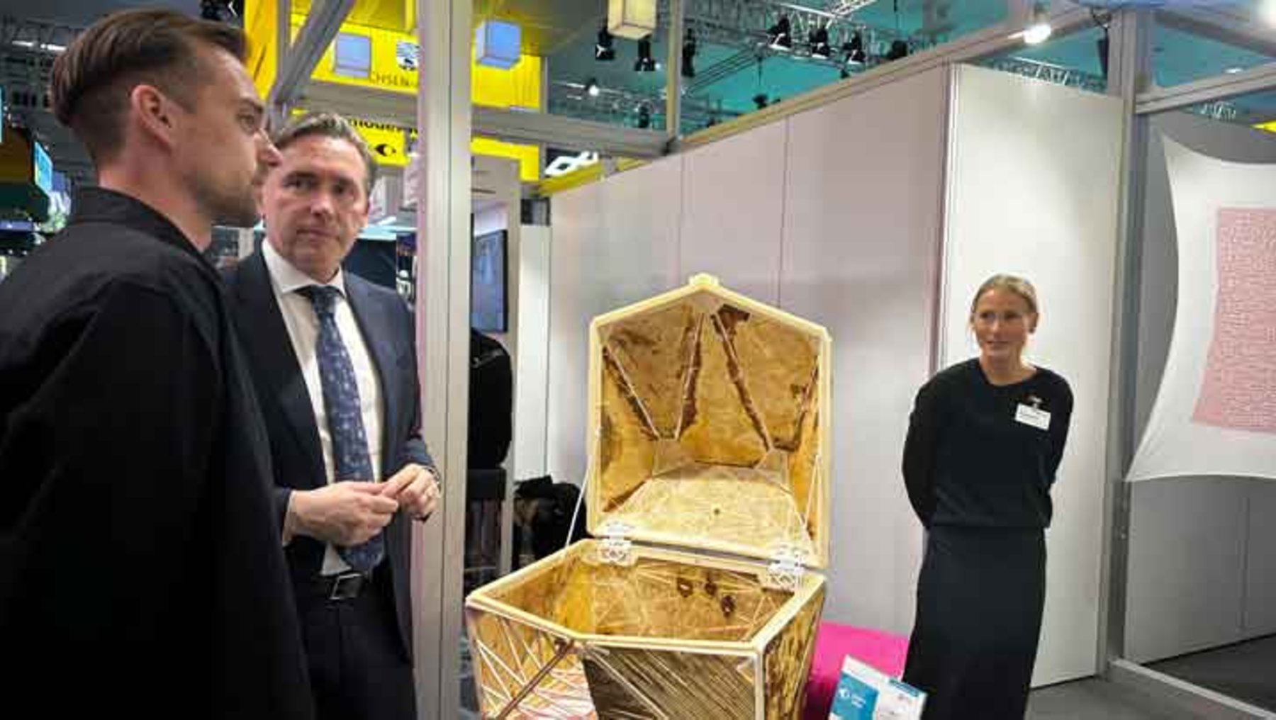Drei Personen auf einer Messe: Zwei Männer in Anzügen betrachten einen großen, offenen, mit Stoff ausgelegten Sarg oder Behälter aus Holz, während eine Frau in schwarzer Kleidung mit Namensschild daneben steht und erklärt.