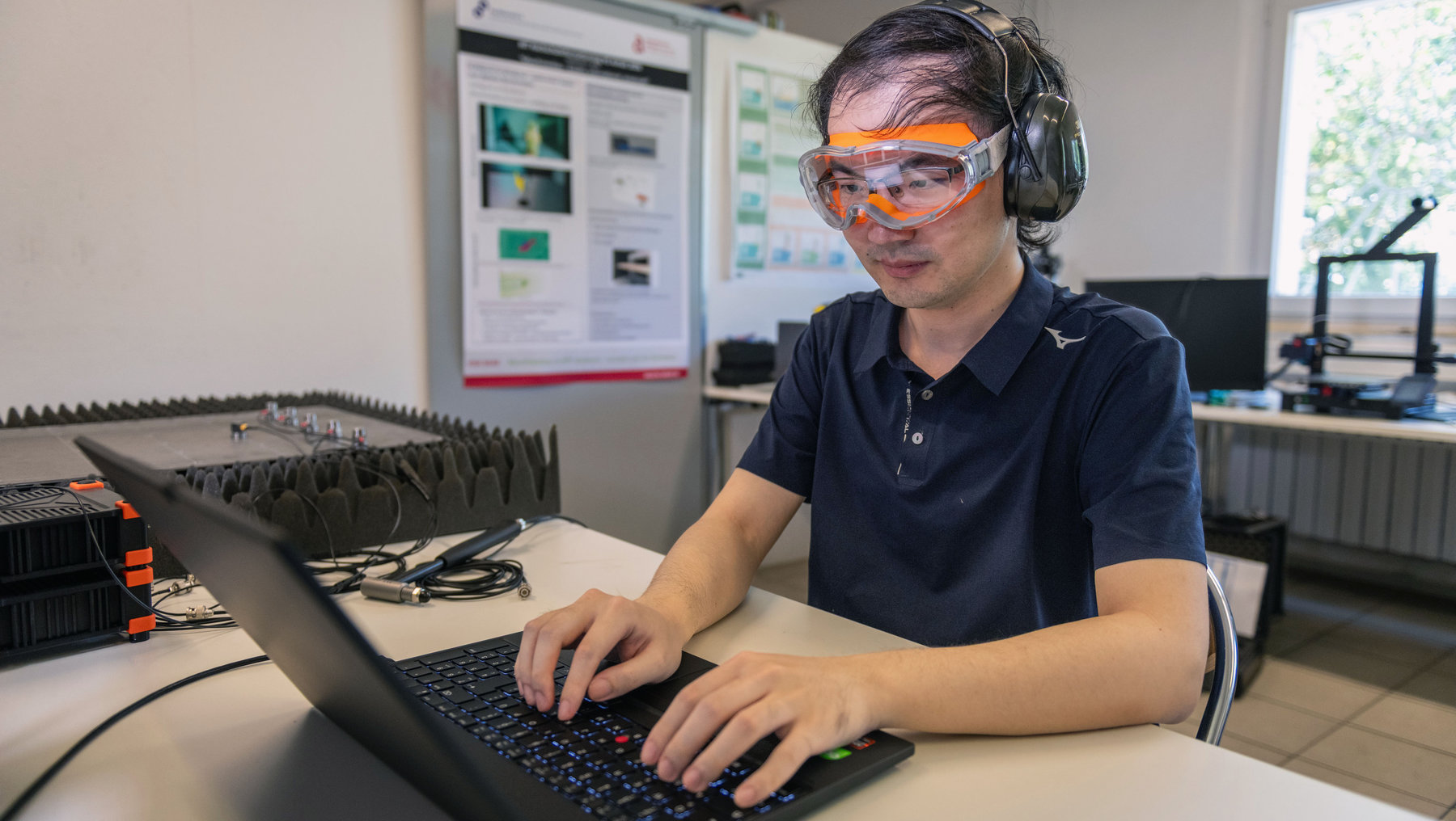 Eine Person sitzt an einem Arbeitsplatz mit Laptop und arbeitet konzentriert. Sie trägt Schutzbrille und Kopfhörer, während vor ihr eine Reihe von Steckplätzen mit elektronischen Bauteilen zu sehen ist. Im Hintergrund hängen Poster mit technischen Darstellungen.