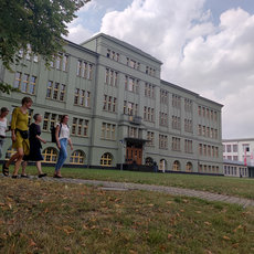 Four people walking together on a grassy area in front of a large, light green, multi-story building with many windows, likely a school or institution. Trees and a clear sky are in the background.