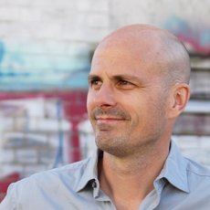 A man with a shaved head and mustache, wearing a light gray button-up shirt, stands in front of a colorful graffiti wall. He is looking off to the side with a calm expression. The graffiti features vibrant colors and abstract shapes.